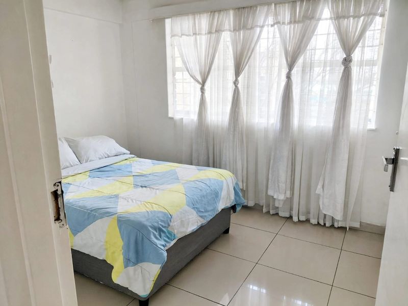 A bright bedroom featuring tiled flooring and a large window fitted with security bars, offering a clean and functional living space.