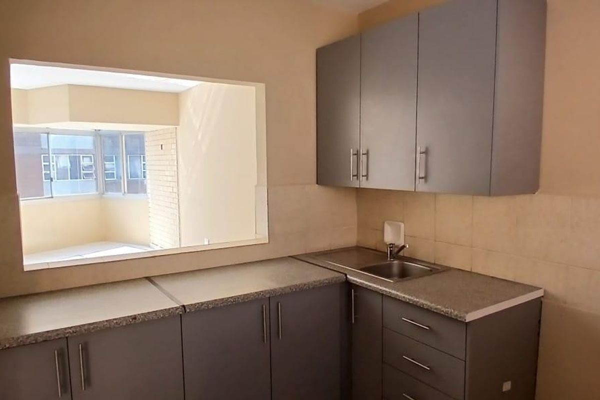 A compact kitchen featuring grey cabinetry, laminate countertops, a stainless steel sink, and a convenient pass-through window looking into the adjacent living area.