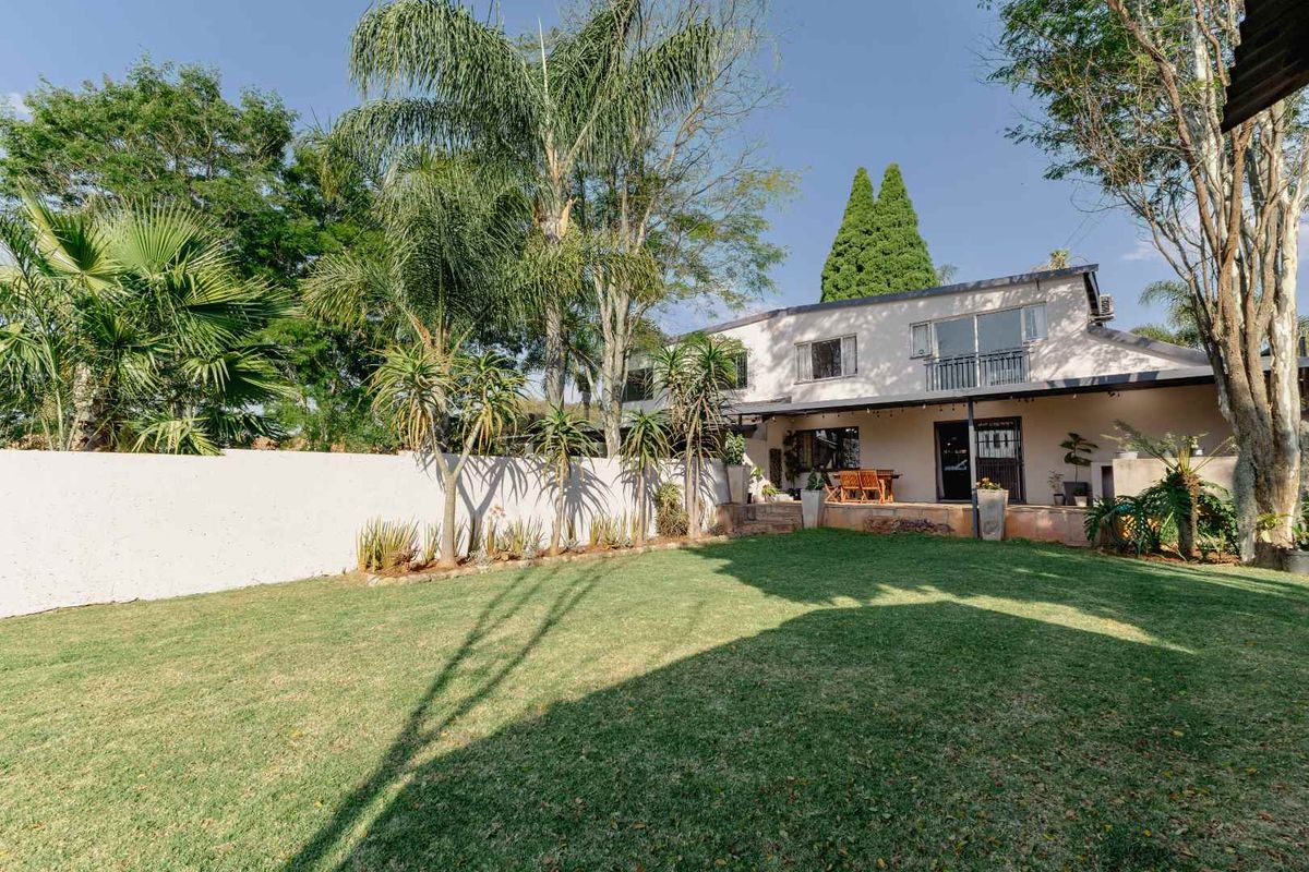 A spacious rear garden featuring a well-maintained lawn, mature trees, a boundary wall, and a covered patio area with an upper-level balcony.