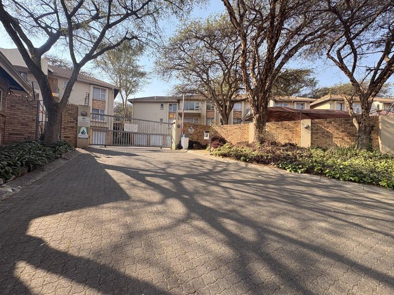 View of the apartment complex entrance featuring a secure gated access point, brick boundary walls, and a paved driveway surrounded by mature trees.
