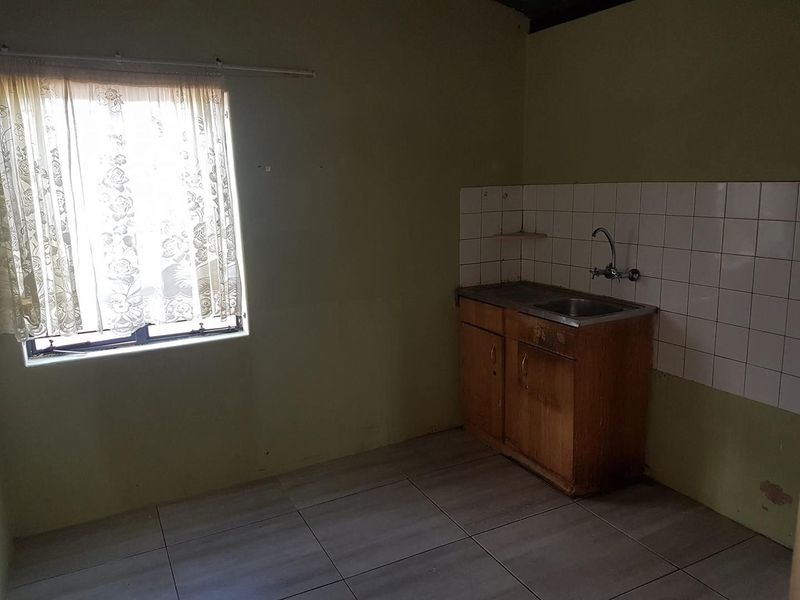 A basic, empty kitchen area featuring tiled flooring, a simple sink unit with tiled splashback, and dated cabinetry in need of refurbishment.