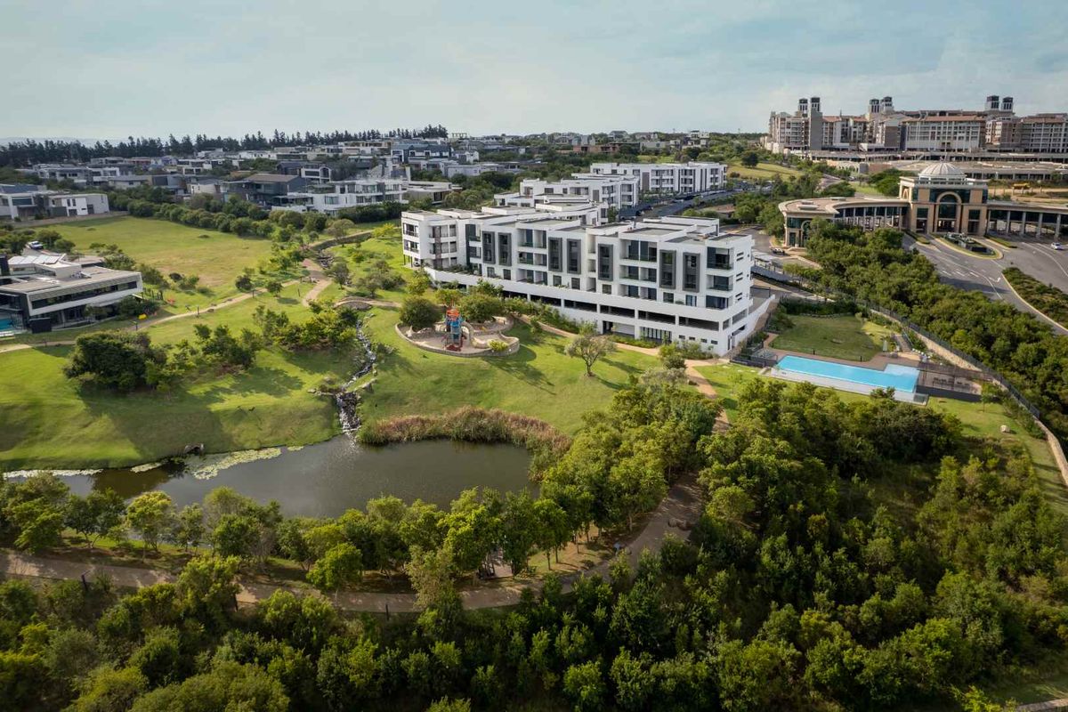 Aerial view of a modern apartment complex featuring landscaped gardens, a swimming pool, and a scenic pond in a well-maintained estate setting.