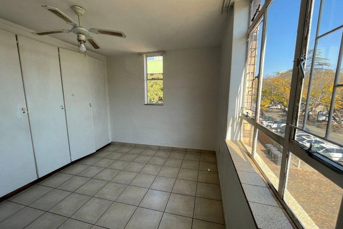 A bright, empty bedroom featuring built-in cupboards, tiled flooring, and large windows providing natural light and an exterior view.
