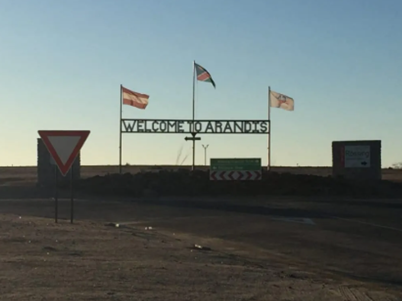 A street-level view of the Arandis town entrance sign featuring flagpoles and directional road signage in a desert landscape setting.