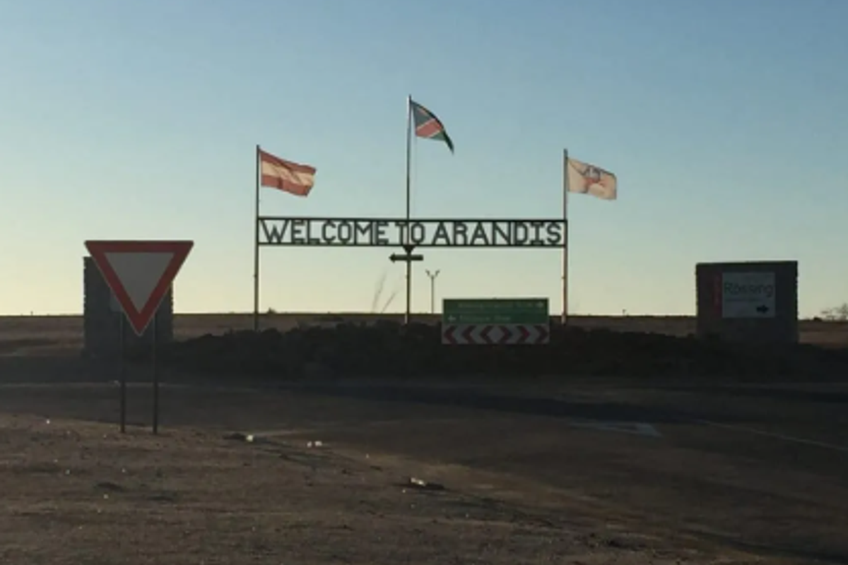 A street-level view of the Arandis town entrance sign featuring flagpoles and directional road signage in a desert landscape setting.