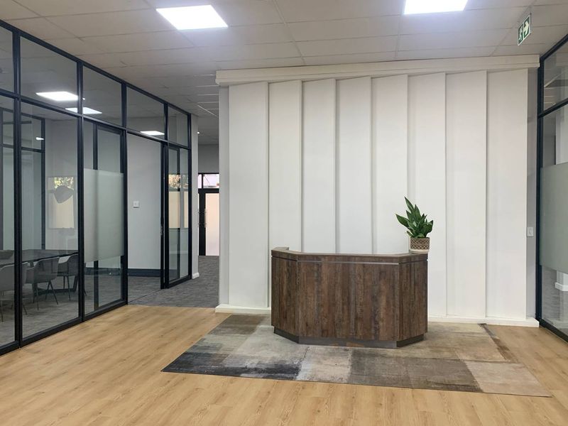 Modern office reception area featuring a wood-finish reception desk, glass-partitioned meeting rooms, and contemporary laminate flooring with recessed ceiling lighting.
