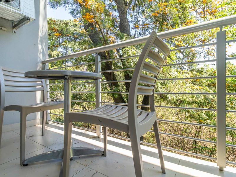 A private balcony featuring tiled flooring, a secure metal balustrade, and a wall-mounted air conditioning unit, overlooking lush greenery.