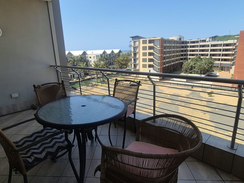 A covered balcony featuring tiled flooring and a stainless steel balustrade, offering an elevated view of the surrounding apartment complex and grounds.