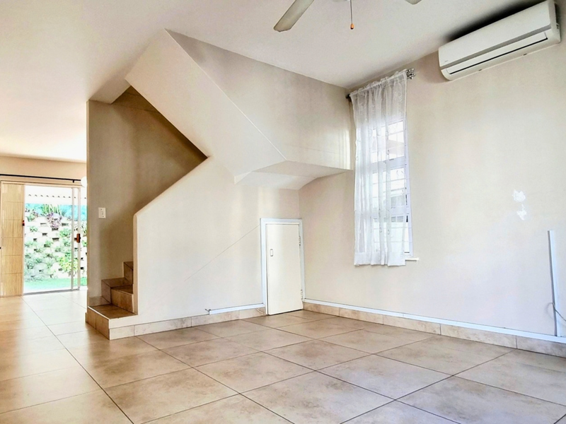 Open-plan living area featuring tiled floors, a staircase with under-stair storage, and an integrated air conditioning unit, leading to a kitchen and outdoor access.
