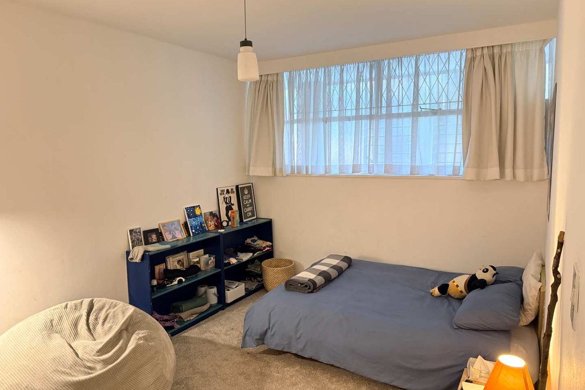 A functional bedroom featuring carpeted flooring, a large window with security bars, and a central pendant light fixture.