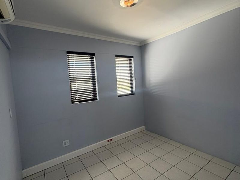 A clean, empty office space featuring tiled flooring, split-system air conditioning, and dual windows with fitted venetian blinds.