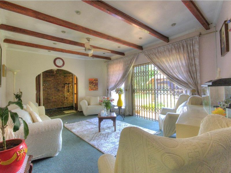 A living room featuring exposed timber ceiling beams, a ceiling fan, and an arched entryway leading to an adjacent space with a sliding security door.