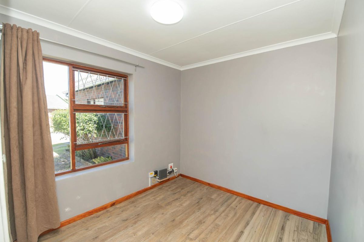 A bright, empty bedroom featuring wood-look laminate flooring, neutral grey walls, and a window with security bars.