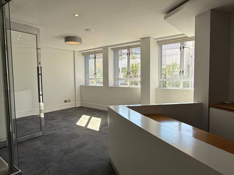 A bright, empty office space featuring a glass partition, built-in reception desk, carpeted flooring, and large windows providing ample natural light.