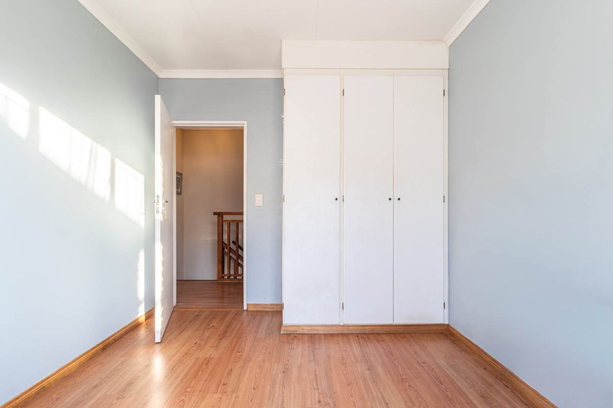 A bright, empty bedroom featuring light blue walls, laminate flooring, and a built-in white wardrobe, with access to a hallway.