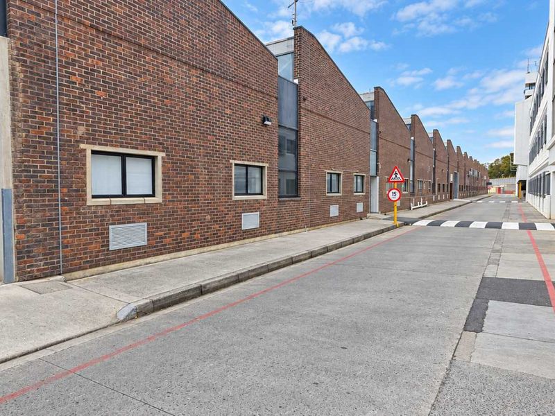 Exterior view of an industrial complex featuring brick facades, concrete driveway access, and roller shutter doors for warehouse entry.