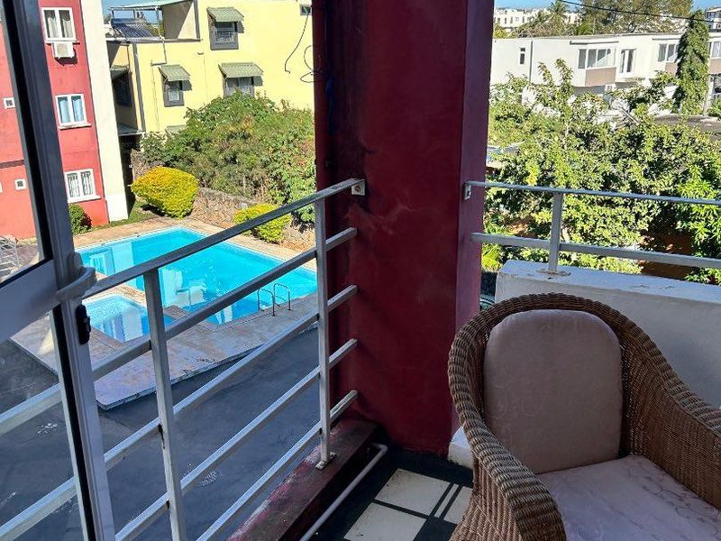 A balcony overlooking a communal swimming pool, featuring a metal balustrade and tiled flooring, requiring maintenance to the structural column and railing.