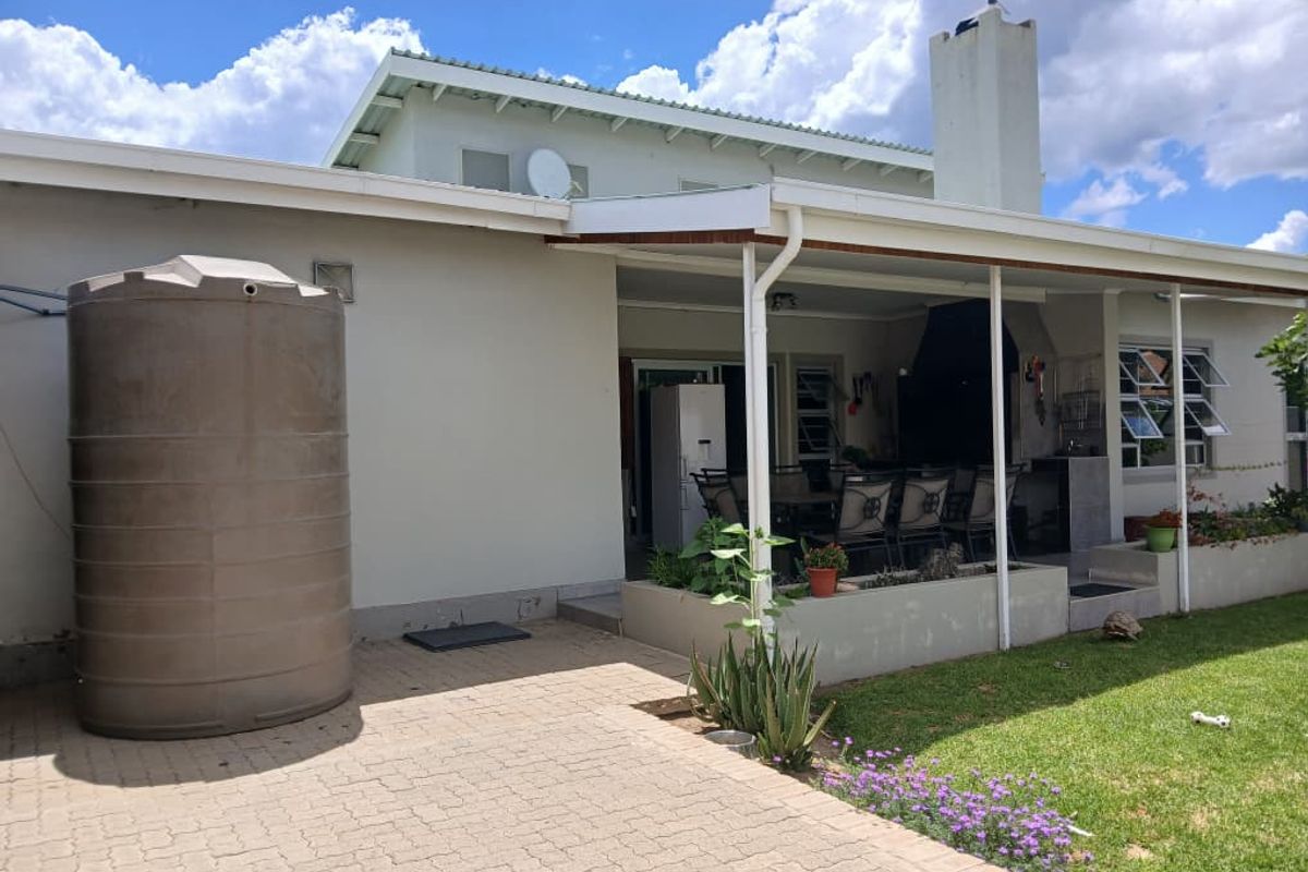 Rear view of the townhouse featuring a covered patio with a built-in braai, paved area, and a large water storage tank in the garden.