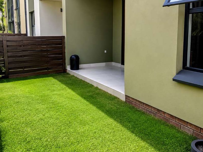 A private ground-floor apartment patio featuring tiled flooring, artificial grass, and a timber privacy fence, providing a low-maintenance outdoor living space.