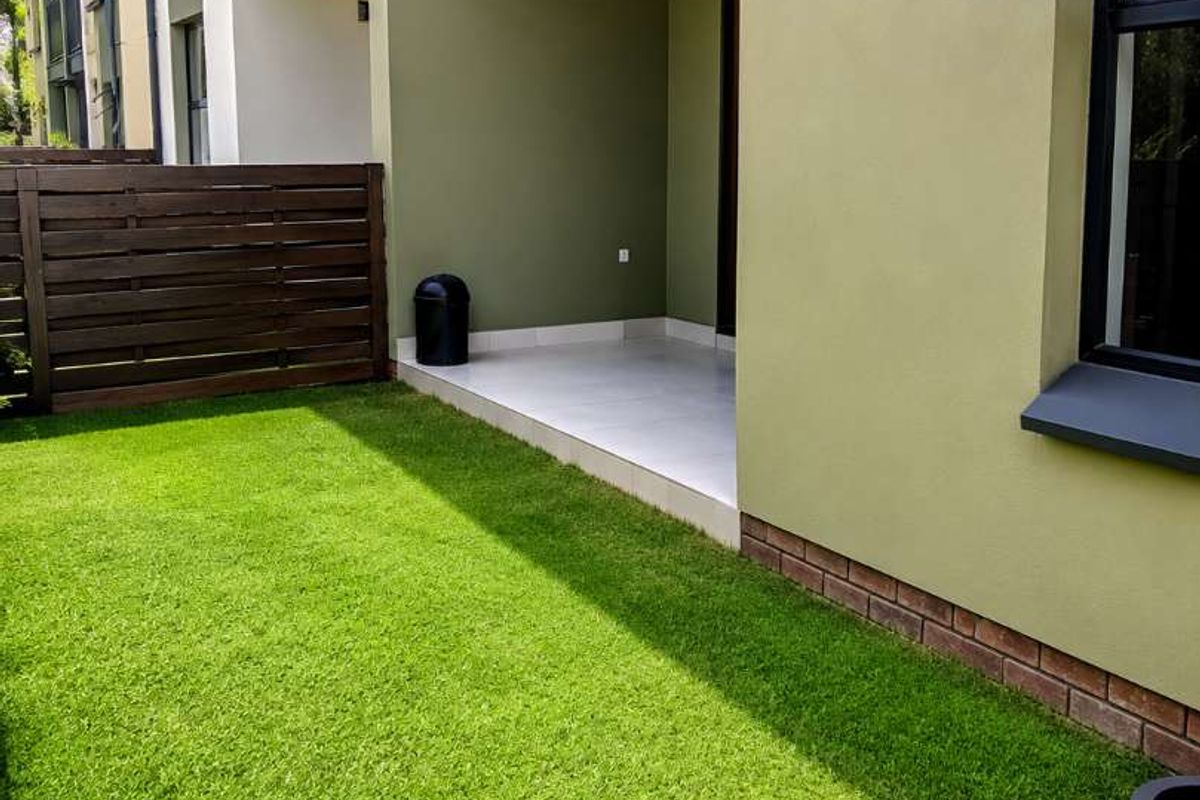 A private ground-floor apartment patio featuring tiled flooring, artificial grass, and a timber privacy fence, providing a low-maintenance outdoor living space.