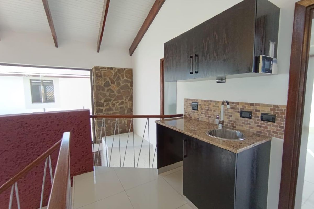 A compact kitchenette area featuring dark cabinetry, granite countertops, a stainless steel sink, and a tiled backsplash, situated near a staircase landing.