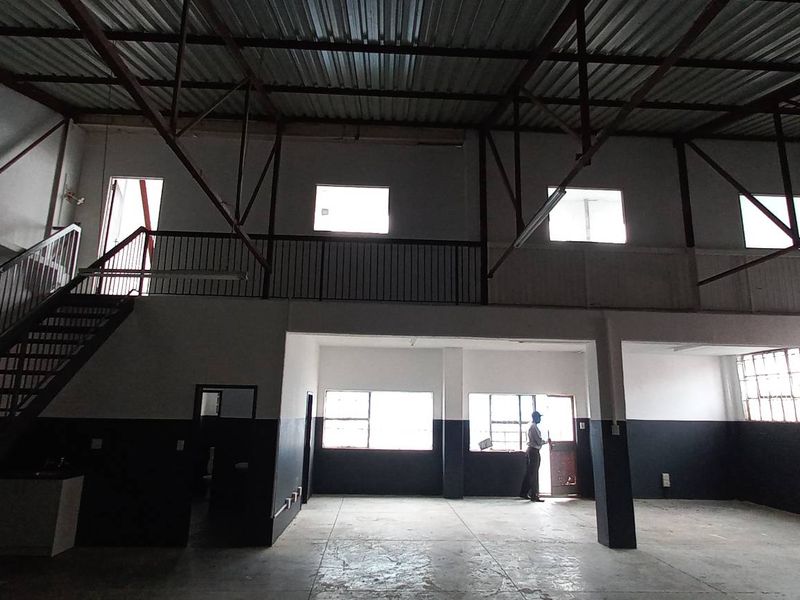 Spacious industrial warehouse unit featuring a mezzanine level, exposed steel roof trusses, concrete flooring, and large windows providing ample natural light.