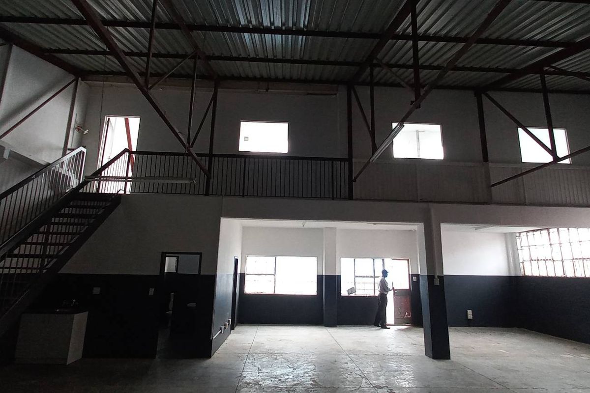 Spacious industrial warehouse unit featuring a mezzanine level, exposed steel roof trusses, concrete flooring, and large windows providing ample natural light.