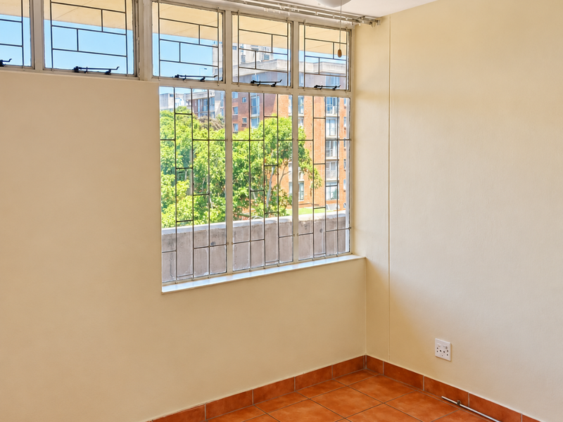An empty bedroom featuring terracotta floor tiles, a ceiling fan, and large windows with security bars overlooking the surrounding neighborhood.