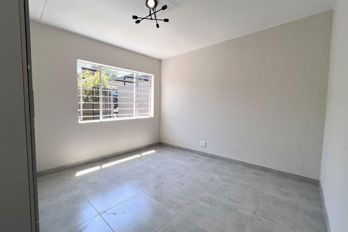 A bright, empty bedroom featuring neutral tiled flooring, a modern ceiling light fixture, and a window fitted with security bars.