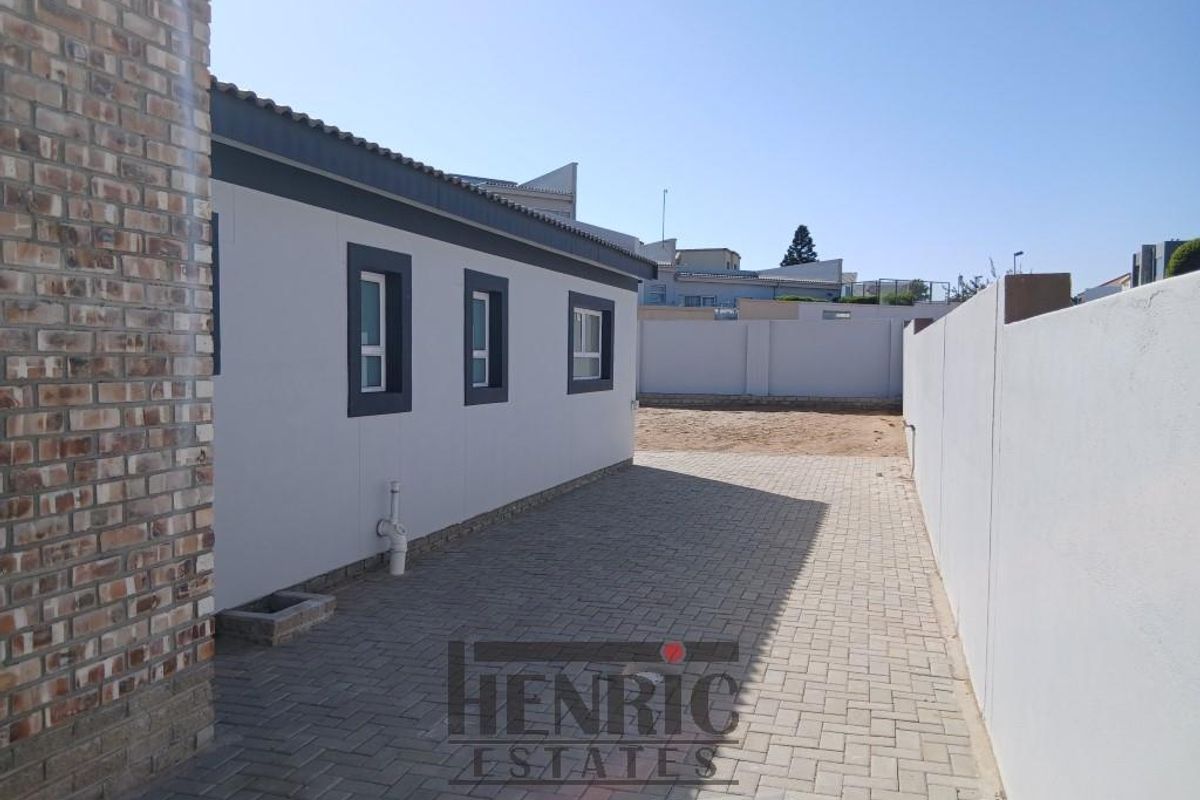 A side exterior view of the property featuring paved driveway access, boundary walls, and modern window frames.