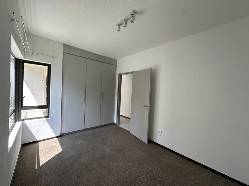 A clean, empty bedroom featuring neutral-toned built-in cupboards, carpeted flooring, and natural light from a large window.