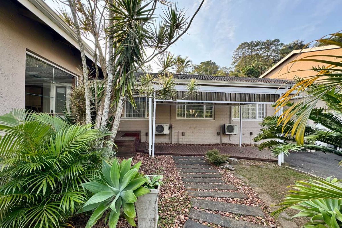 A rear exterior view featuring a covered timber deck, stone pathway, and established garden landscaping adjacent to the house.