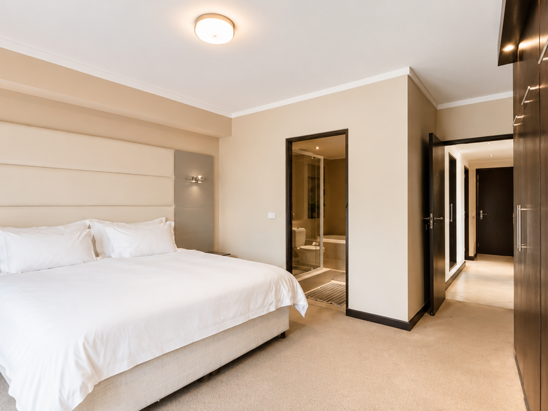 Spacious bedroom featuring a large upholstered headboard, integrated wall lighting, and extensive built-in wardrobe storage with direct access to an en-suite bathroom.