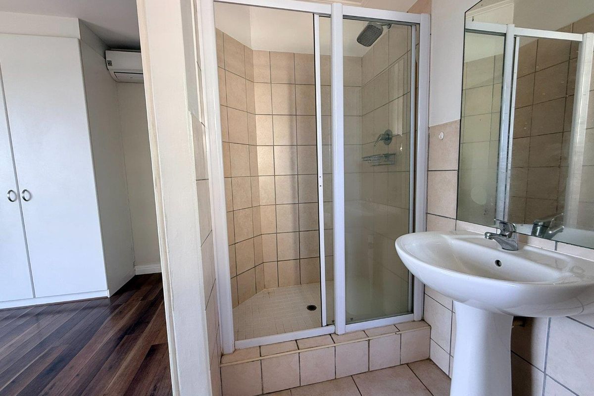 A functional bathroom featuring a tiled shower enclosure, pedestal basin, and wall-mounted mirror, adjacent to a bedroom area with built-in storage.