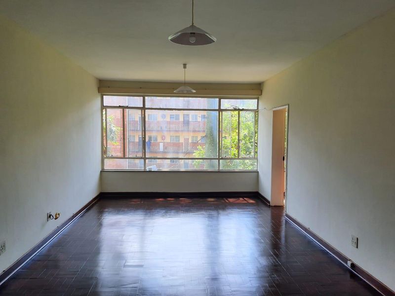 An empty living room featuring dark parquet flooring, large windows providing natural light, and simple pendant lighting, ready for personalization.