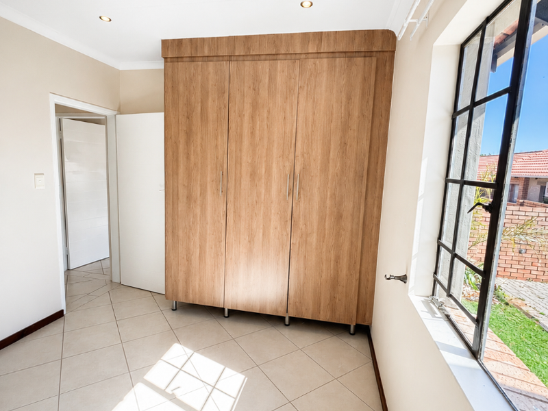 A bright, empty bedroom featuring neutral tiled flooring, built-in wooden cupboards, and a large steel-framed window overlooking the exterior.