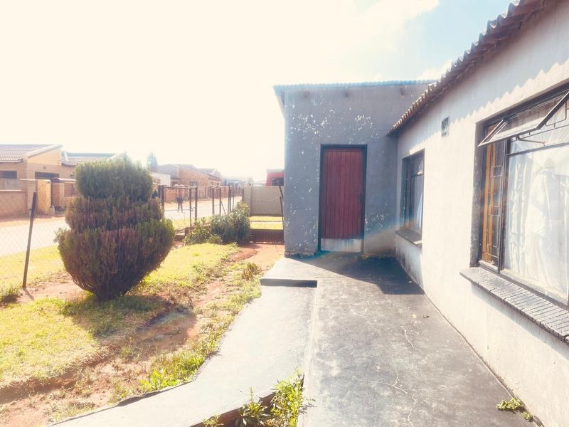 Front exterior view of the house featuring a concrete walkway, metal window frames, and a corrugated roof, showing signs of weathered exterior paint.