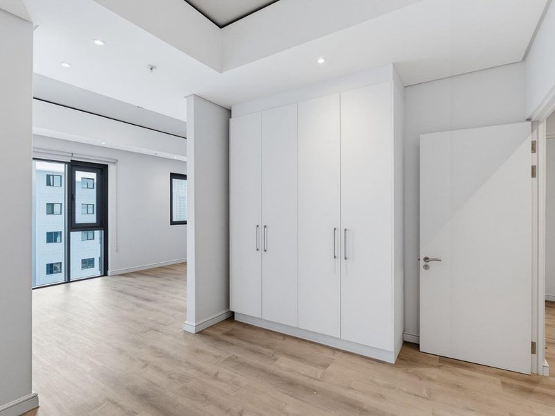 A bright, modern bedroom featuring neutral finishes, built-in cupboards, laminate flooring, and recessed ceiling lighting, offering a clean and spacious living environment.