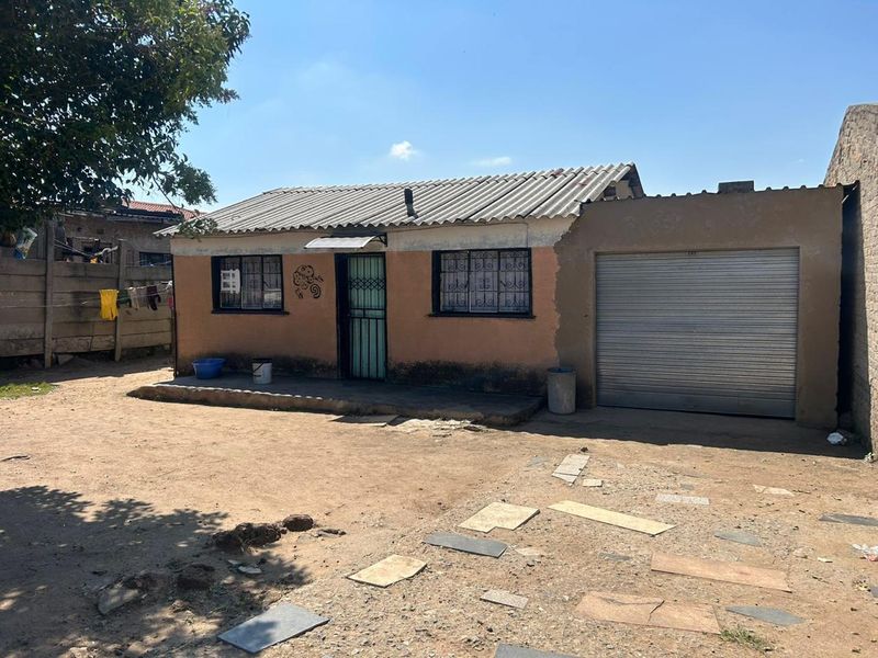 Single-story residential property featuring a corrugated roof, security bars on windows, and an integrated roller garage door with an unpaved front yard.