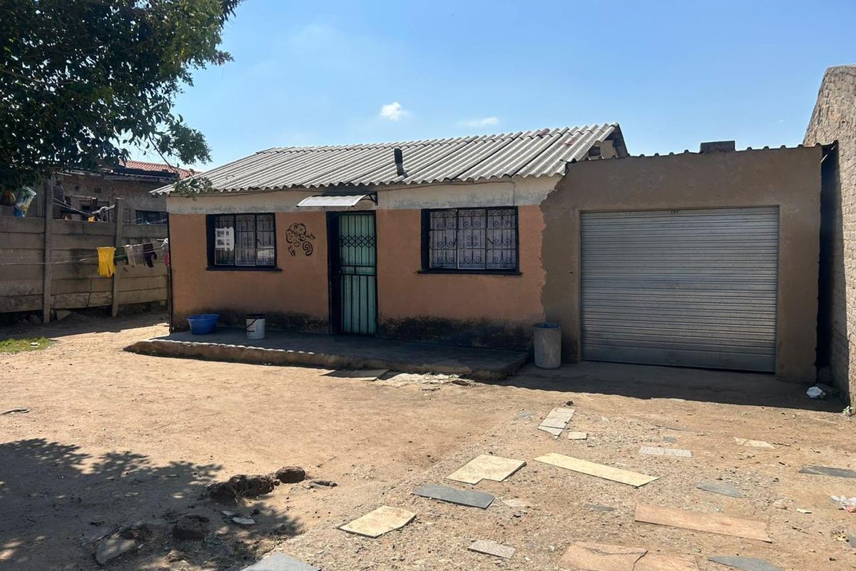 Single-story residential property featuring a corrugated roof, security bars on windows, and an integrated roller garage door with an unpaved front yard.