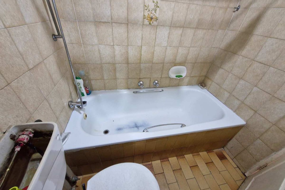 A dated bathroom featuring a built-in bathtub with shower attachment and a toilet with an exposed cistern, requiring significant renovation and modernization.