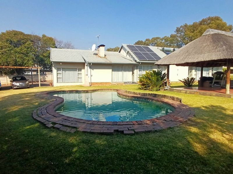 Rear garden view featuring a swimming pool with brick coping, a thatched lapa, and a main house equipped with solar panels.