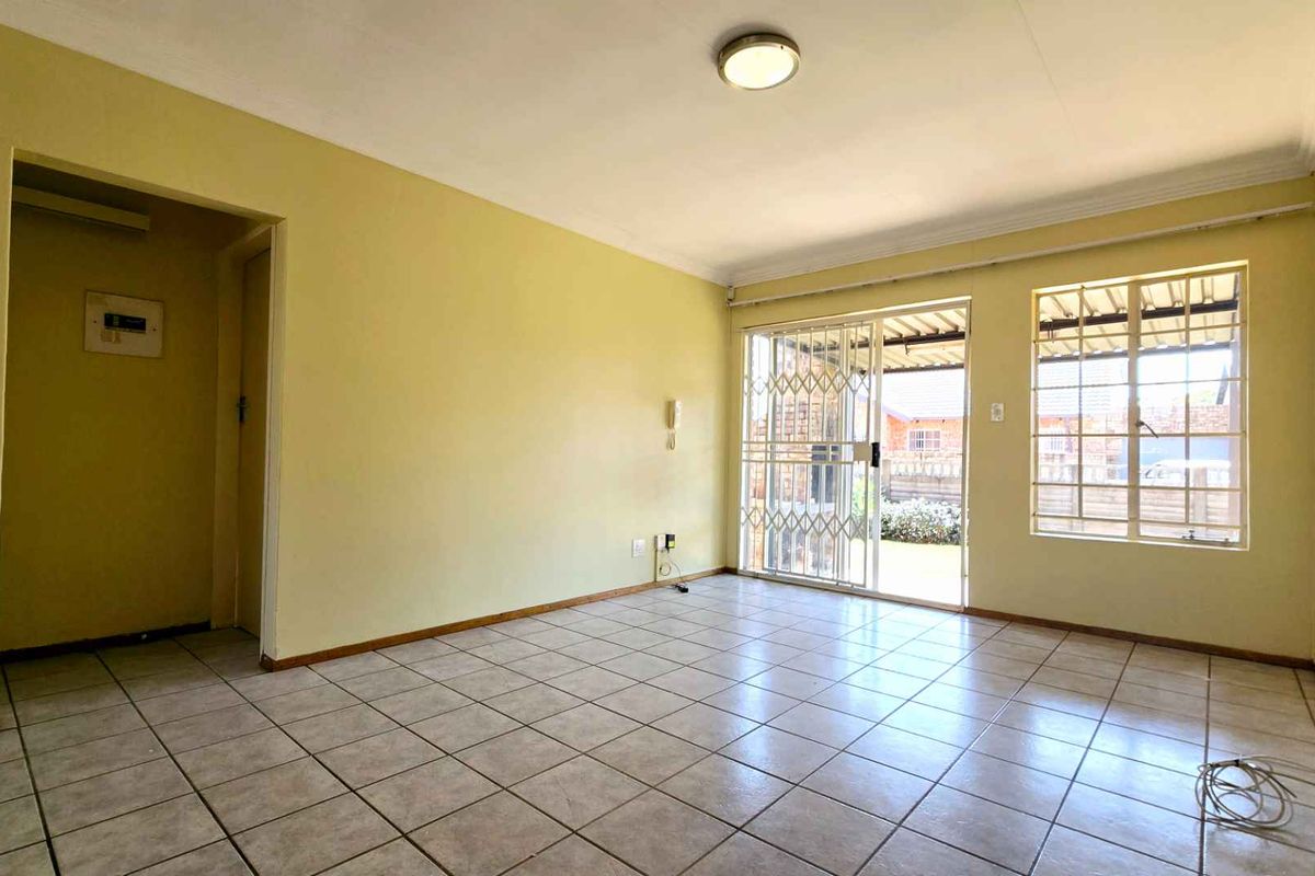A spacious, empty living area featuring tiled flooring, large windows with security bars, and a sliding door leading to the exterior.