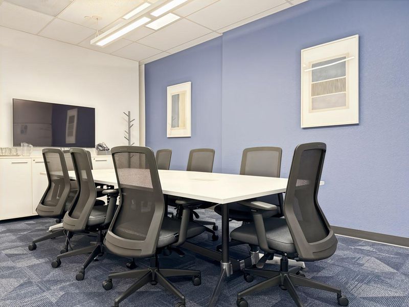 A professional office meeting room featuring a large conference table, built-in storage cabinetry, suspended ceiling with recessed lighting, and blue carpet tiles.