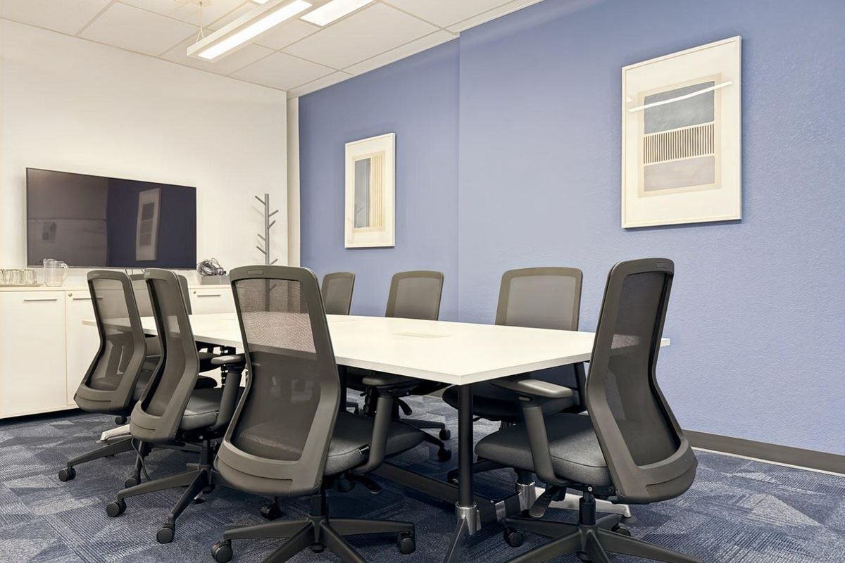 A professional office meeting room featuring a large conference table, built-in storage cabinetry, suspended ceiling with recessed lighting, and blue carpet tiles.