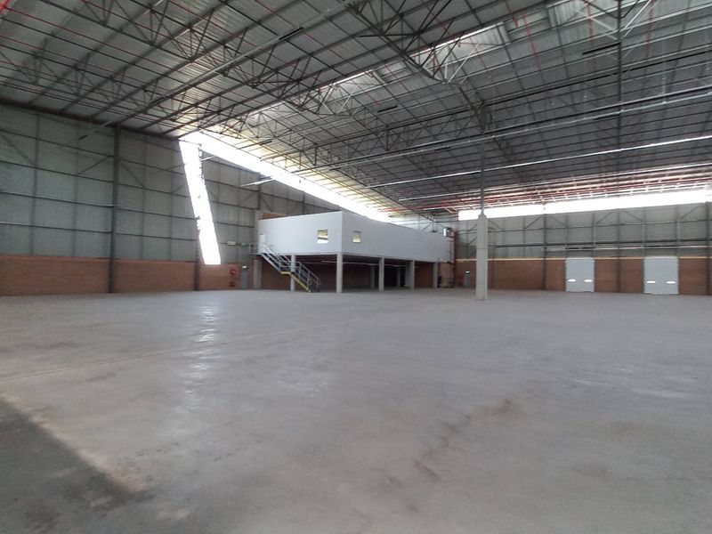 Spacious industrial warehouse featuring high ceilings with exposed steel trusses, concrete flooring, a mezzanine office level, and multiple roller shutter doors.