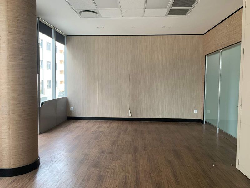 A vacant office space featuring wood-look flooring, a frosted glass partition, and large windows, providing a bright and professional work environment.