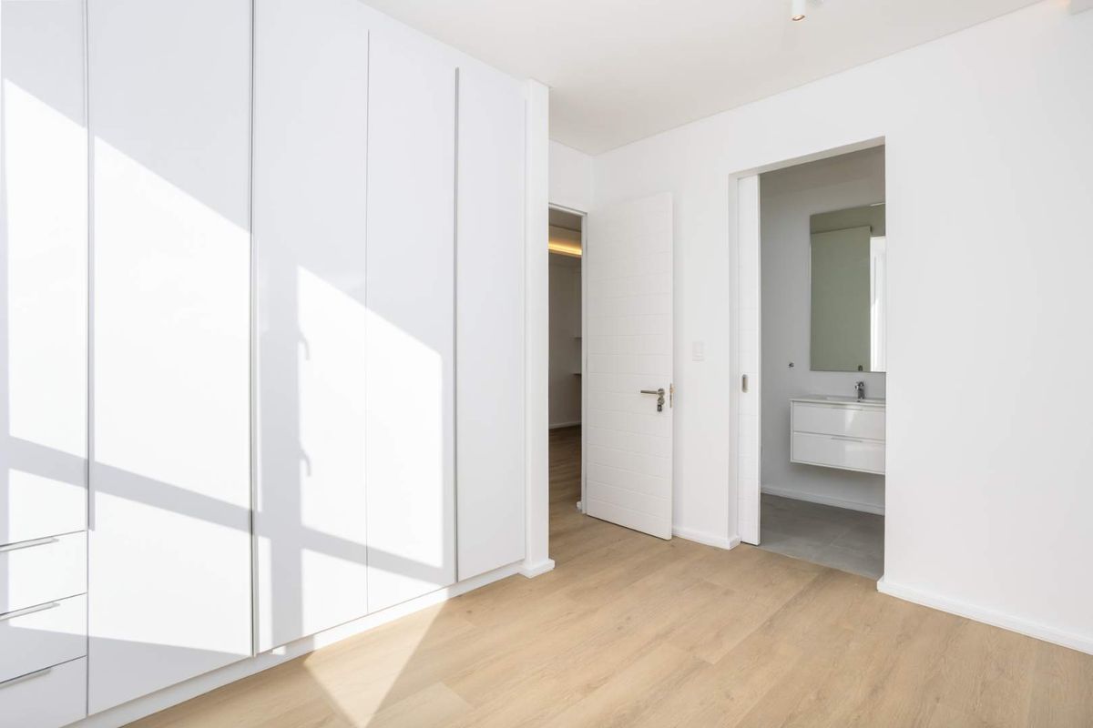 A bright bedroom featuring built-in floor-to-ceiling wardrobes, light wood-look flooring, and direct access to an en-suite bathroom with a modern vanity.