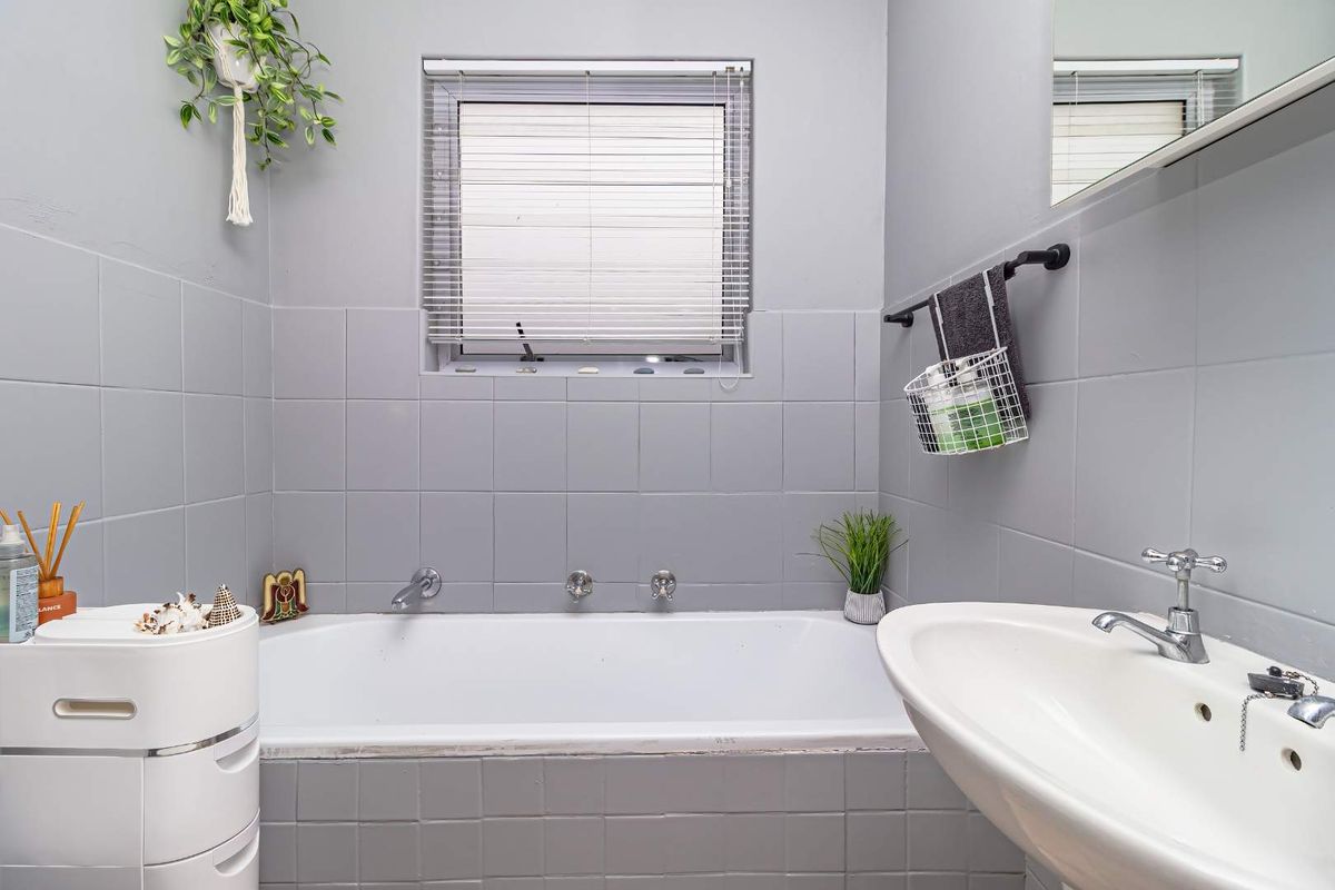 A clean, functional bathroom featuring a built-in bathtub, wall-mounted basin, and neutral grey wall tiling with a window for natural light.