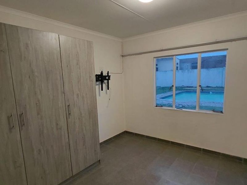 A clean, empty bedroom featuring built-in cupboards, tiled flooring, and a window view overlooking the swimming pool area.